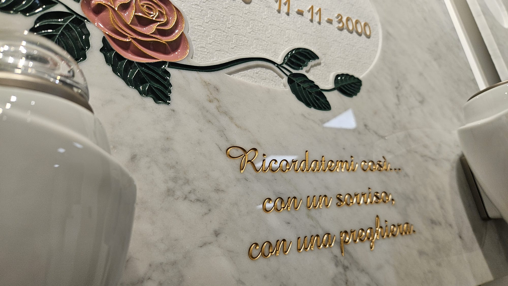 Lapide commemorativa in marmo chiaro con decorazione floreale a rosa rosa e foglie verdi. Incisa in lettere dorate la dedica: ‘Ricordatemi così… con un sorriso, con una preghiera".