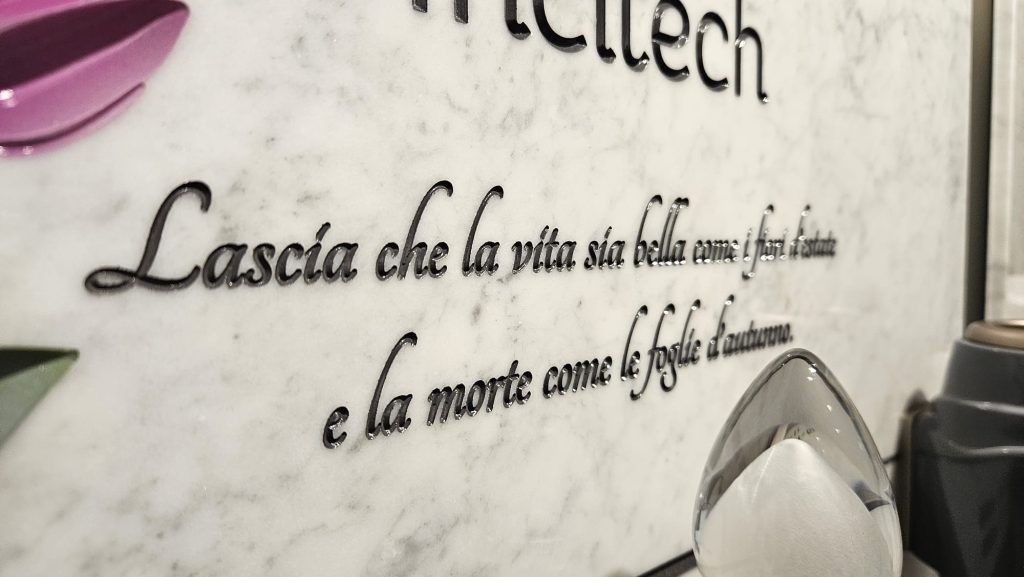 Dettaglio di lapide commemorativa in marmo bianco con incisione in rilievo lucida. La dedica per lapide recita: "Lascia che la vita sia bella come i fiori d’estate e la morte come le foglie d’autunno”.